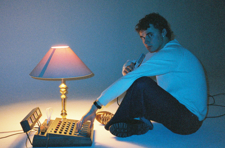 Musician Maybe Hugo sitting on floor with blue lighting, holding microphone next to table lamp and mixing console in studio setting.